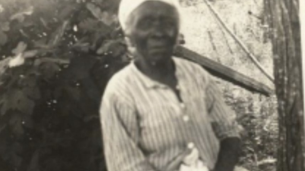 black and white portrait photograph of Elizabeth Burney, an elder African-American woman. she is seated in front of a bush wearing a white kerchief, a striped button-down shirt, and a shite printed skirt.